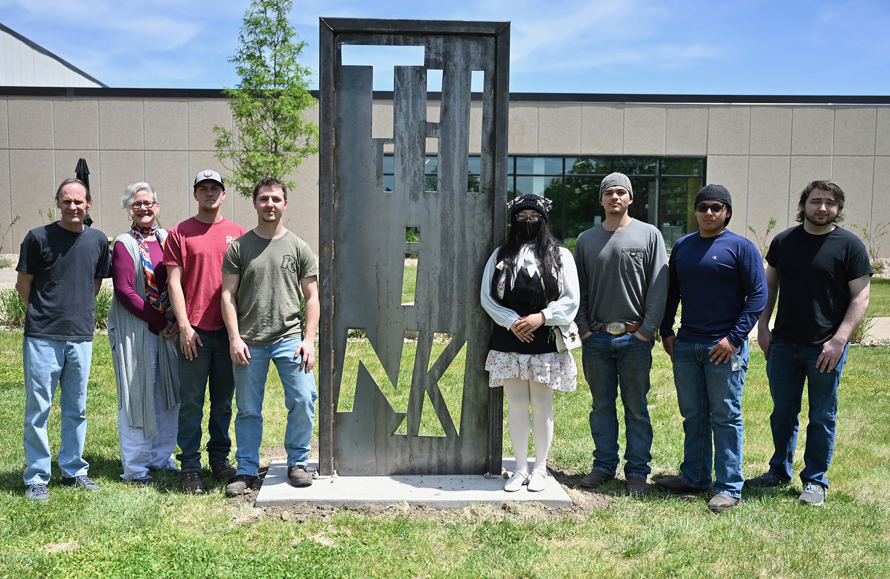 It’s Why You’re Here by KPaw Law Moo. Pictured (L to R): Tim Castle, Nancy Adams, Noah Gallentine, Carter Wagner, KPaw Law Moo, Hector Torres, Alvaro Castro Sical, and Dalton Black. Group of eight people standing on a campus lawn around a tall, rectangular metal sculpture with cut-out letters, positioned in front of a college building and a small tree on a sunny day.